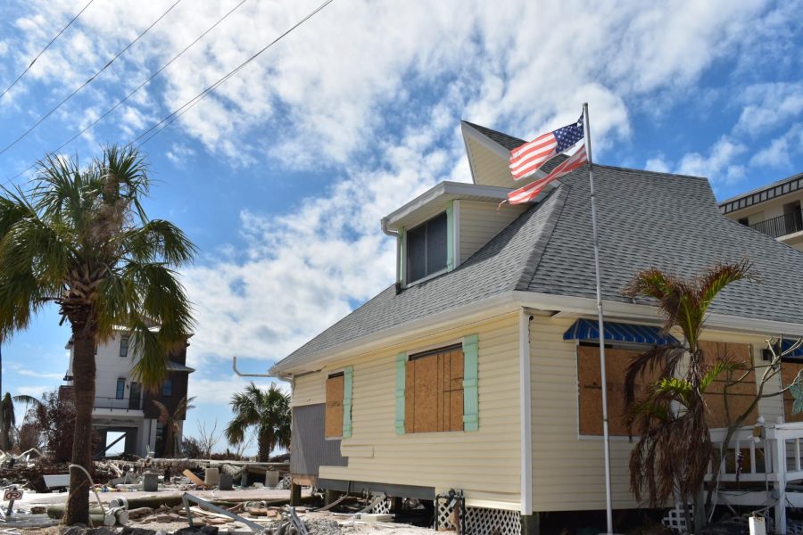 A house stands behind a torn United States flag with boarded windows and doors having some damage to the exterior.